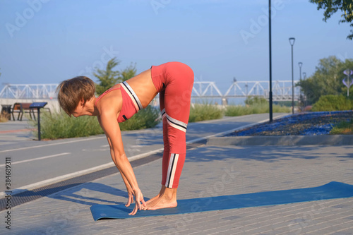 Young attractive woman in red sportswear practices yoga exercises outdoors.