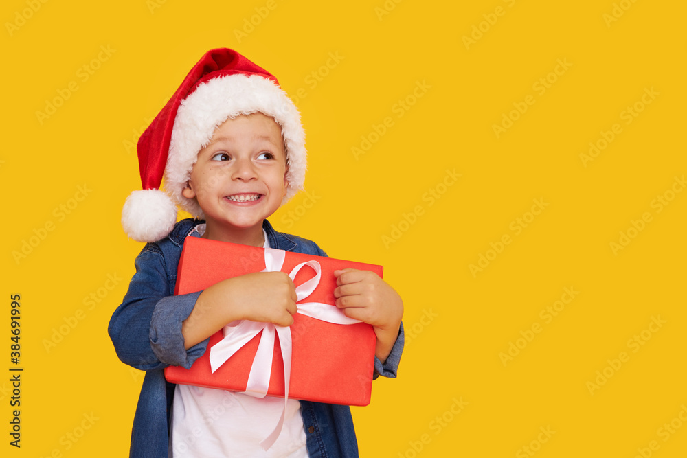 Portrait of a satisfied little child boy in christmas Santa hat. laughing isolated over yellow background. Holds a gift box. Preparing for the New Year holidays
