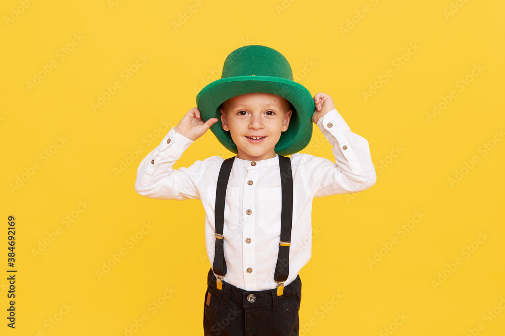 Smiling little child boy in green leprechaun hat on yellow background. St. Patrick Day celebration. Funny face