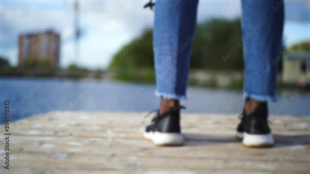 Blurred image. Back for inscriptions. Girl black sneakers, blue jeans entered pier near river, lake in park, downtown. Forest and house in the background. Tourist destination. Exploring new places. Blurred image. Back for inscriptions. Girl black sneakers, blue jeans entered pier near river, lake in park, downtown. Forest and house in the background. Tourist destination. Exploring new places.