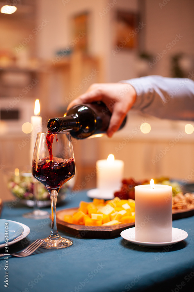 Close up of guy pouring wine in glass during romantic dinner with wife in dining room. Young man pouring red wine in wife glass. Romantic caucasian happy couple sitting at the table celebrating .
