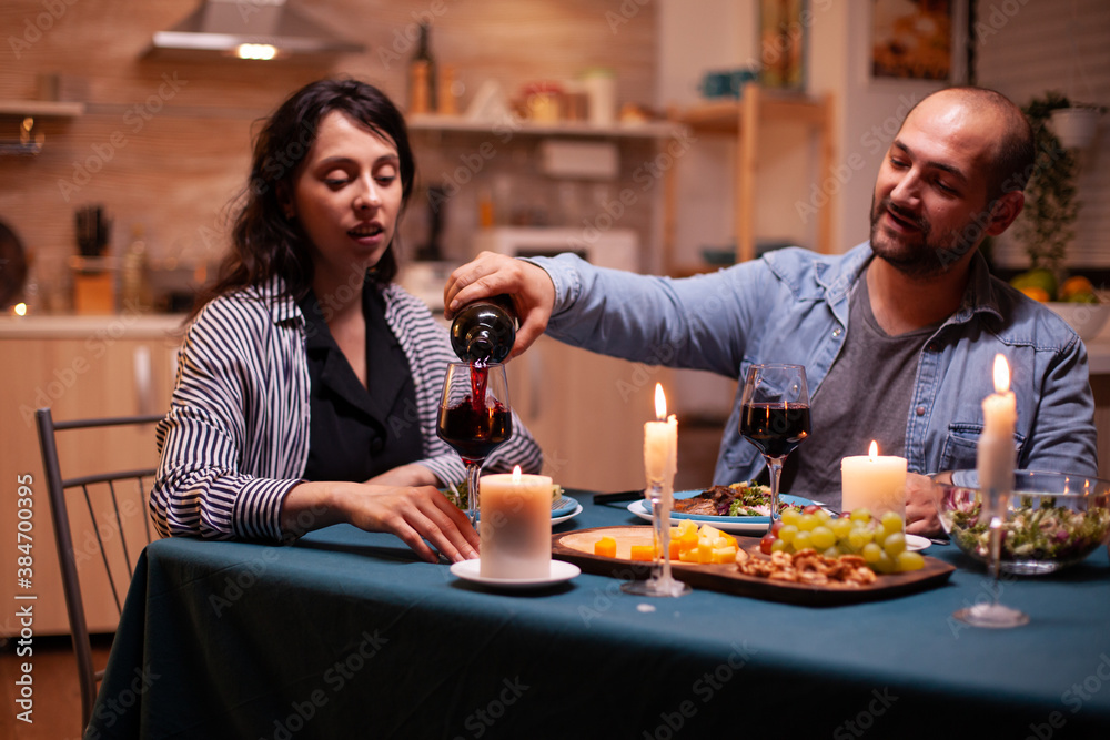 Boyfriend celebrating relationship with wife in dining room. young man pouring red wine in wife glass. Romantic caucasian happy couple sitting at the table celebrating .