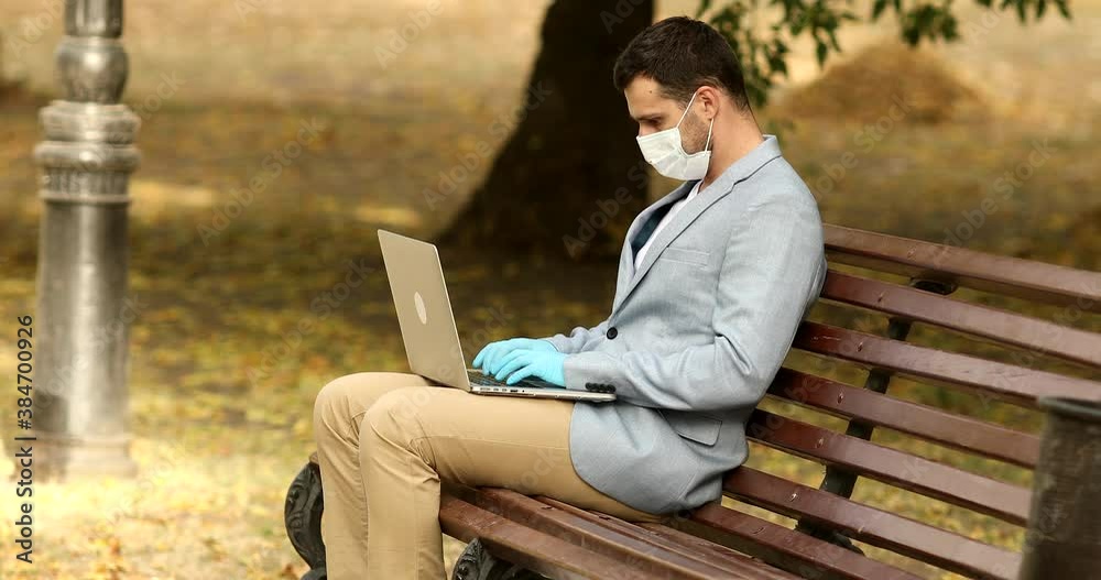 Young businessman with face mask working outdoors on bench in city, using laptop. Covid-19. Coronavirus. Young businessman with face mask working outdoors on bench in city, using laptop. Covid-19. Coronavirus.