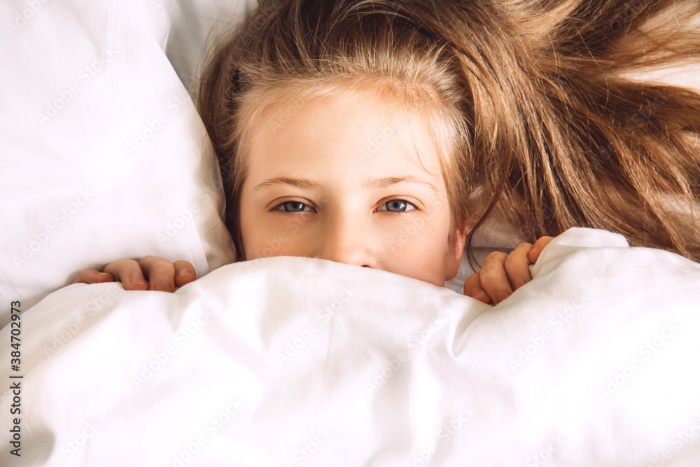 Child hiding under duvet in bed. Morning routine. Indulge in the bedroom in the morning. Girl in bed look out from under white covers. Selective focus Child hiding under duvet in bed. Morning routine. Indulge in the bedroom in the morning. Girl in bed look out from under white covers. Selective focus