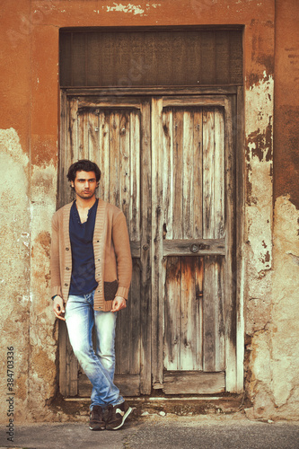 Handsome young man is leaning against the wall near the door of an old house entrance. A ruined wooden door.