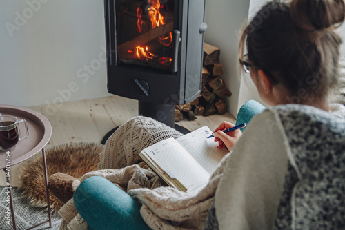 Young woman  write in notebook sitting in armchair by fireplace photo