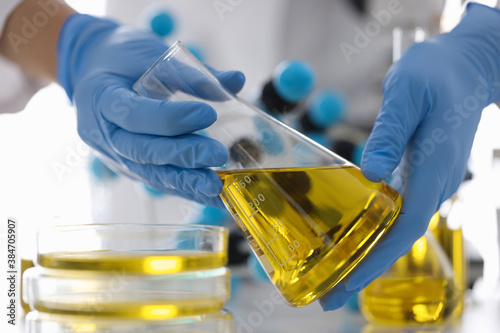 Hands in rubber gloves hold flask with yellow liquid in chemical laboratory closeup. Quality control in oil refining industry concept.