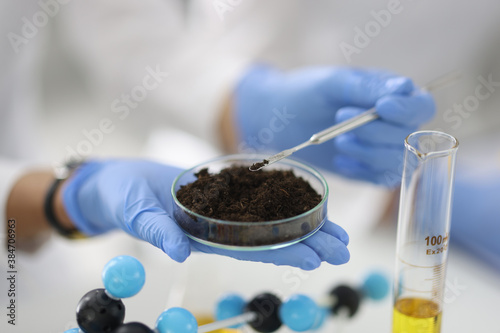 Glass cup with soil sample stands on hand in rubber glove in chemical laboratory closeup. Biochemical analysis of soils concept.