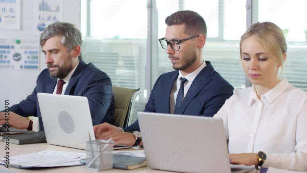 Medium shot of serious businessmen and businesswoman in formalwear sitting at desk and working on laptops Medium shot of serious businessmen and businesswoman in formalwear sitting at desk and working on laptops