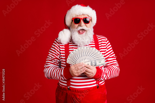 Photo of pensioner old man grey beard hold cash fan play spend money expensive products wear santa x-mas costume suspenders spectacles striped shirt cap isolated red color background © deagreez