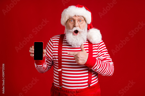 Photo of retired old man grey beard shock hold telephone direct finger screen empty space wear santa x-mas costume suspenders spectacles striped shirt cap isolated red color background © deagreez