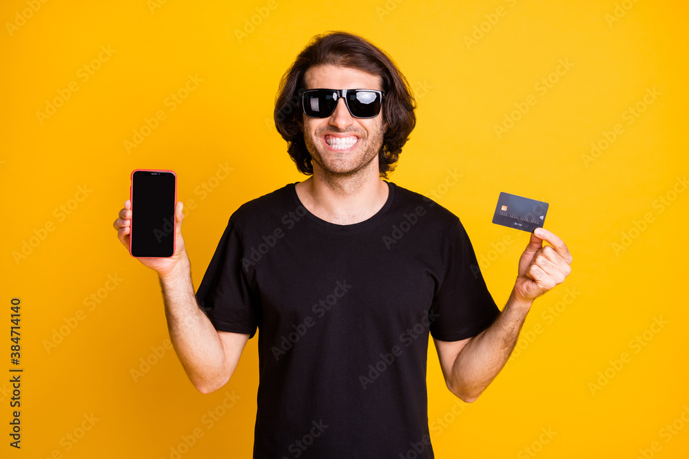 Young man hold debit card telephone empty space toothy smile wear black t-shirt sunglass isolated yellow color background Young man hold debit card telephone empty space toothy smile wear black t-shirt sunglass isolated yellow color background