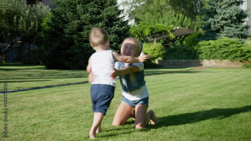 summer holiday, cheerful girl without teeth catches in hands her small brother and lies down on a green grass while walking in the park summer holiday, cheerful girl without teeth catches in hands her small brother and lies down on a green grass while walking in the park