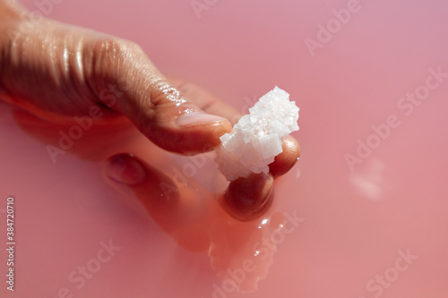 Hand fingers holding white salt crystal in pink vibrant salty water sunny close-up. Spa procedures in natural resort