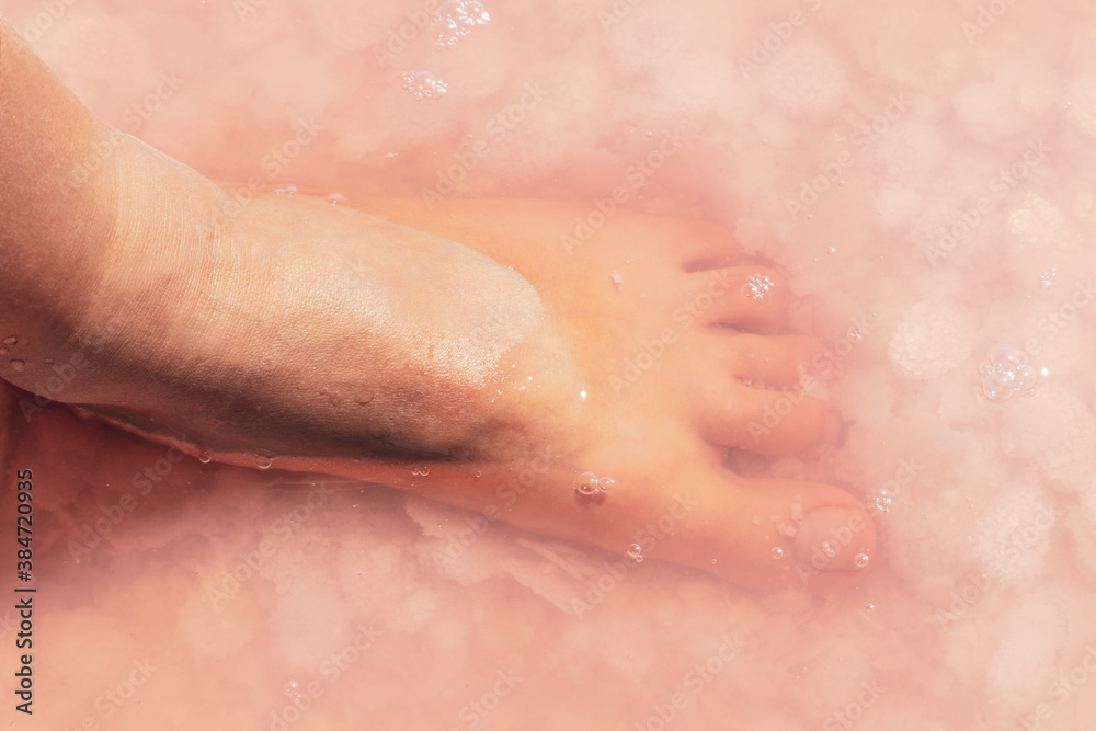 Fototapeta premium Woman leg foot close-up in pink salt flakes top view under pink water surface. Healthy spa procedures