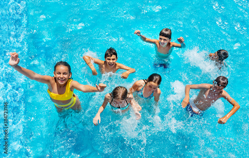 Group of many kids jump and play in swimming pool, splashing, lift hands smile view from above