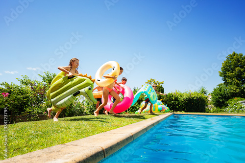 Group of many young kids run and jump into the swimming pool holding inflatable buoy toys diving in the water