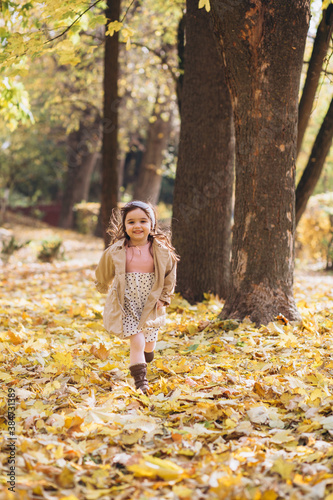 Happy and beautiful little girl in a beige coat runs holding a maple leaf in the autumn park