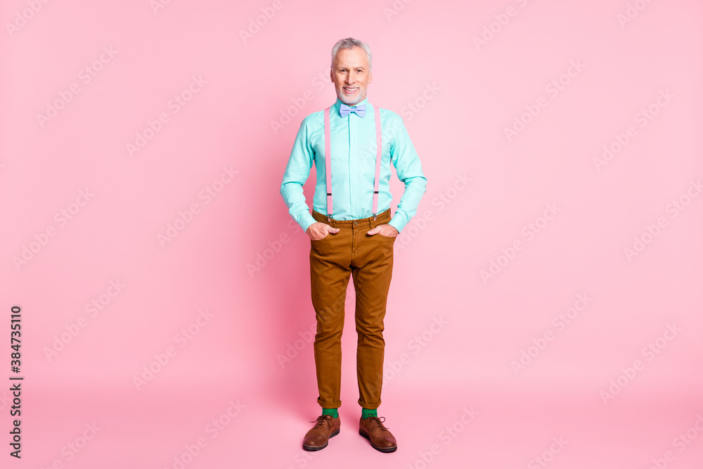 Full length body size portrait of man wearing bowtie suspenders brown pants keeping hands in pockets isolated on pink color background Full length body size portrait of man wearing bowtie suspenders brown pants keeping hands in pockets isolated on pink color background