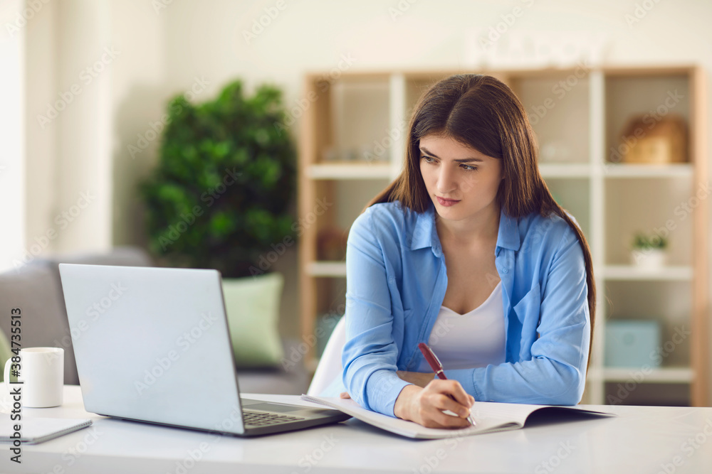 Young female student writing in notebook while watching online lecture sitting at desk at home Young female student writing in notebook while watching online lecture sitting at desk at home