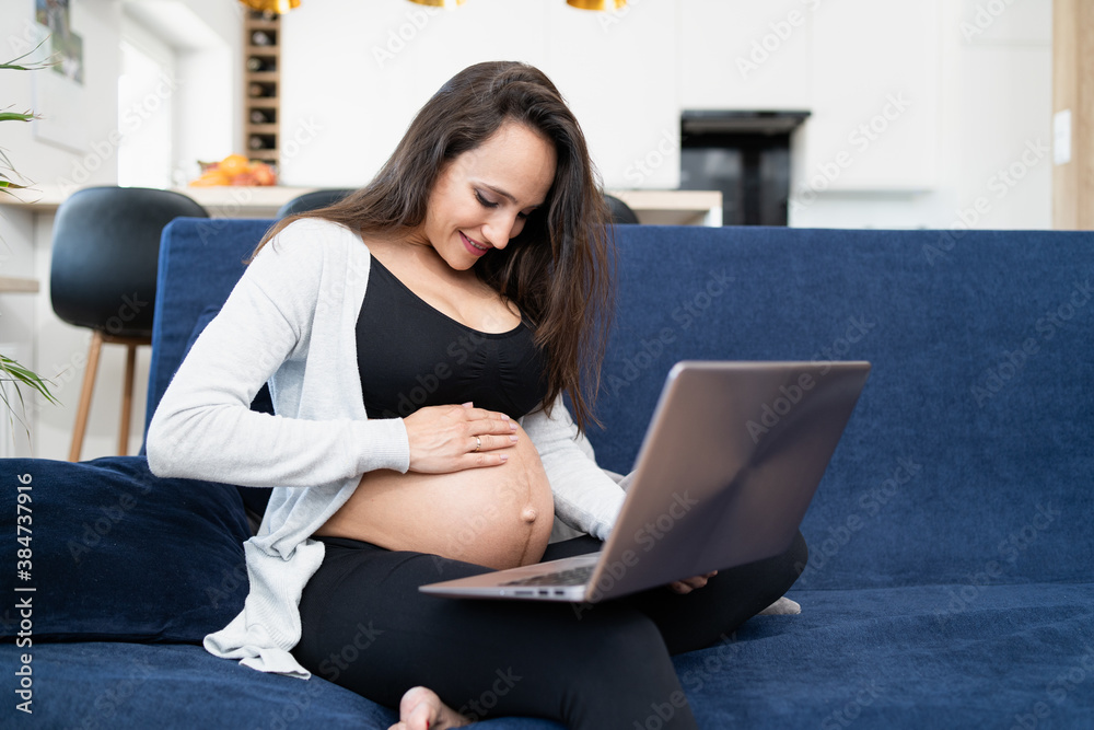 A pregnant brunettre woman is sitting on a sofa in the living room, touching her belly and talking with husband on a video chat. She is wearing sports clothes. The kitchen is visible in the background A pregnant brunettre woman is sitting on a sofa in the living room, touching her belly and talking with husband on a video chat. She is wearing sports clothes. The kitchen is visible in the background