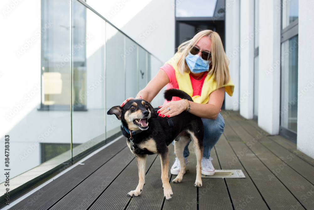Young woman at home on a terrace with protective mask. Hugging her cute black dog. Corona virus Covid-19 concept. Quarantine theme. woman is not leaving home because of a pandemic. Lonely in illness.