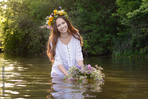 A beautiful young smiling girl with flowing dark hair and a wreath on her head is standing in a river and holding a wreath of flowers