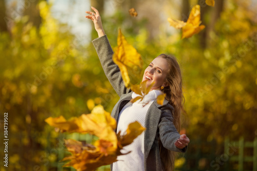 Beautiful happy woman with light brown hair smiles and raises her hands. Leaf fall in the autumn park. Attractive young woman in a gray coat and white sweater. Yellow foliage, Fall season. Autumn mood