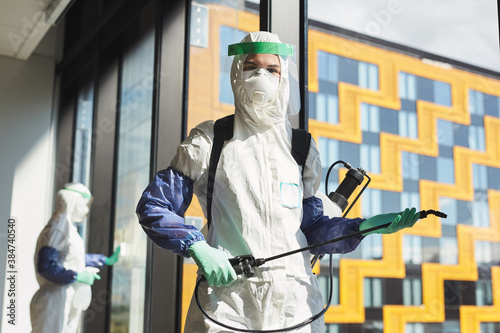 Waist up portrait of female worker wearing hazmat suit and holding disinfecting equipment looking at camera while standing in office, copy space
