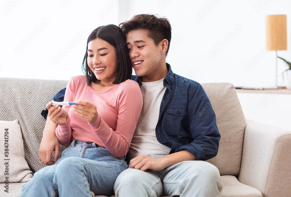 Joyful Asian Couple Holding Positive Pregnancy Test Sitting At Home Joyful Asian Couple Holding Positive Pregnancy Test Sitting At Home