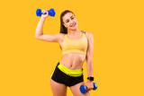 fitness woman in yellow and black sportswear makes pumping hands exercise with dumbbells. sport concept photo. isolated on yellow background. copy space