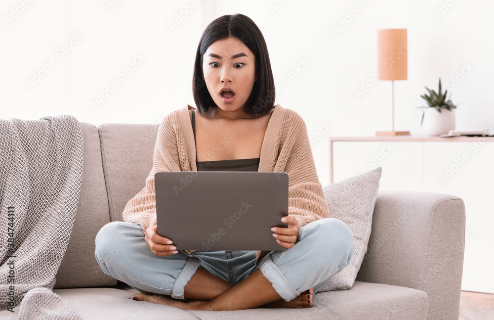 Surprised asian woman sitting on couch, using laptop