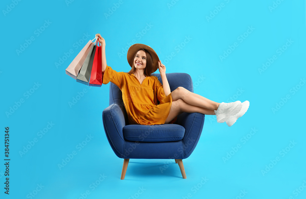 Beautiful young woman with paper shopping bags in armchair on light blue background