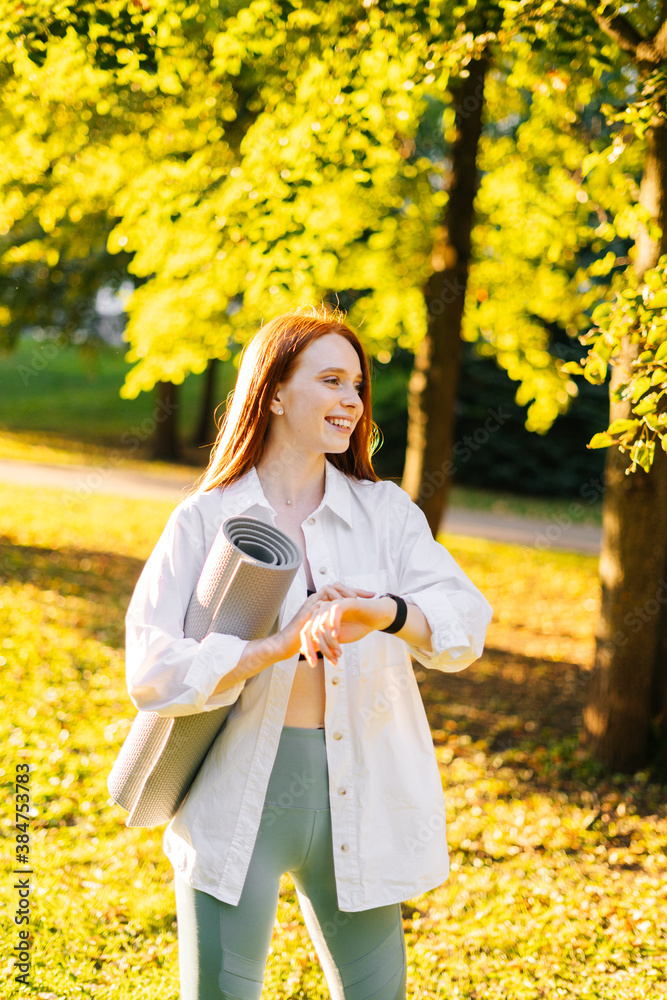 Cheerful happy red-haired young woman using sports smart watch and holding yoga mat in hand while standing at city park in sunny summer morning. Cheerful happy red-haired young woman using sports smart watch and holding yoga mat in hand while standing at city park in sunny summer morning.