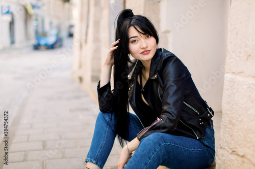 Portrait of young beautiful Asian lady in jeans pants and black leather jacket, sitting on the stone stairs near the old building in city. City lifestyle. Copy space for text.