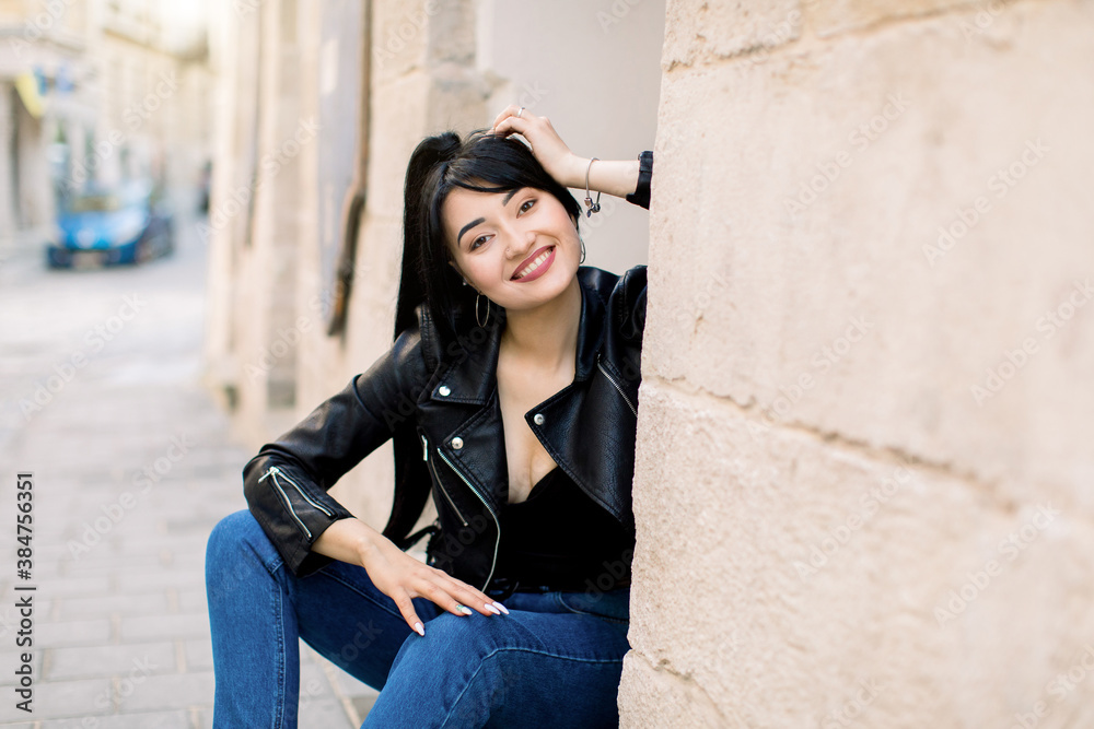 Street portrait of young beautiful cheerful smiling Asian woman with dark ponytail hairstyle, wearing stylish leather jacket and denim jeans, sitting near old building in European city