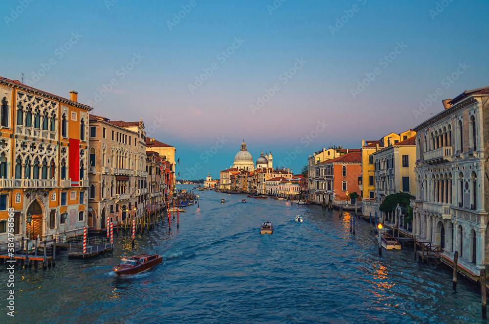 Fototapeta premium Venice cityscape with Grand Canal waterway. Buildings with evening lights along Grand Canal. Santa Maria della Salute Roman Catholic church on Punta della Dogana at twilight. Veneto Region, Italy.