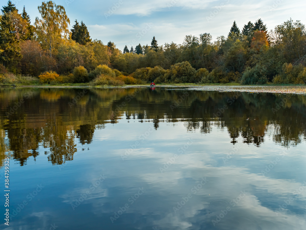 View of the calm water of the lake with the reflection of the autumn forest. A rowing boat is barely visible in the distance