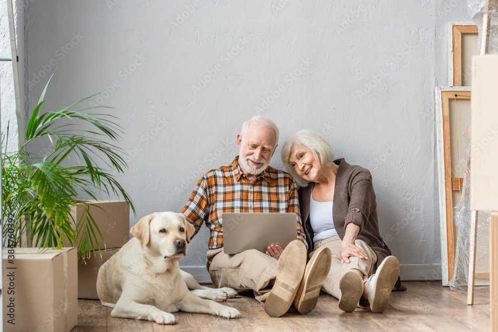 Φωτογραφία happy senior couple using laptop sitting on floor and husband petting dog lying
