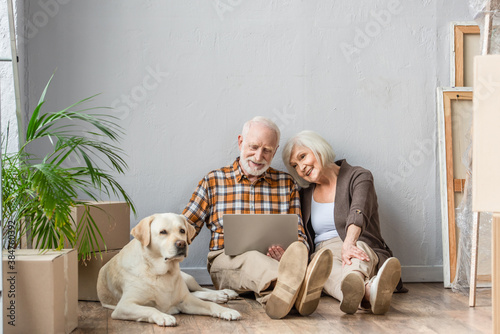 happy senior couple using laptop sitting on floor and husband petting dog lying near