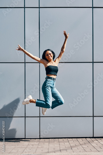 Fototapeta Naklejka Na Ścianę i Meble -  Portrait of cheerful positive black girl jumping in the air on wall street background