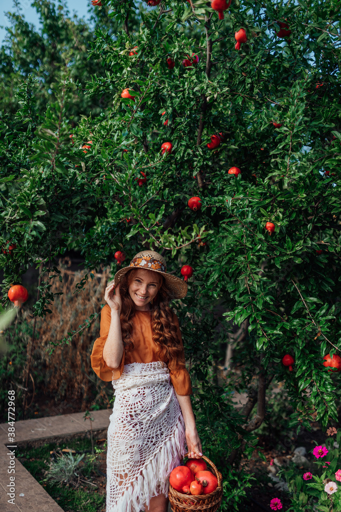 Red pomegranate garden with a happy redhead girl in a hat in an orange dress and white shawl. Woman with big red fruits in .fruit basket, with an amazing smile Red pomegranate garden with a happy redhead girl in a hat in an orange dress and white shawl. Woman with big red fruits in .fruit basket, with an amazing smile