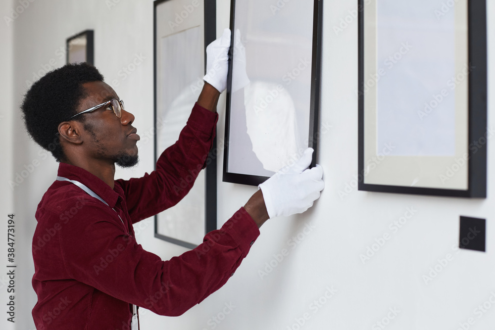 Side view at African-American man hanging frames on wall while planning art gallery or exhibition, copy space
