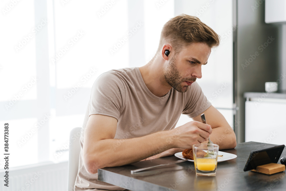 Young man in earphones looking at cellphone while having breakfast at home Young man in earphones looking at cellphone while having breakfast at home