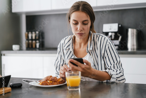 Young caucasian woman holding cellphone while having breakfast at home