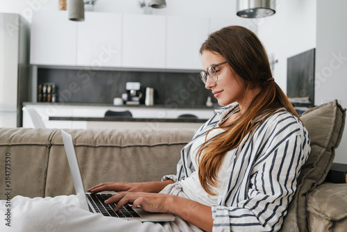 Young caucasian happy woman smiling and using laptop while sitting on sofa at home