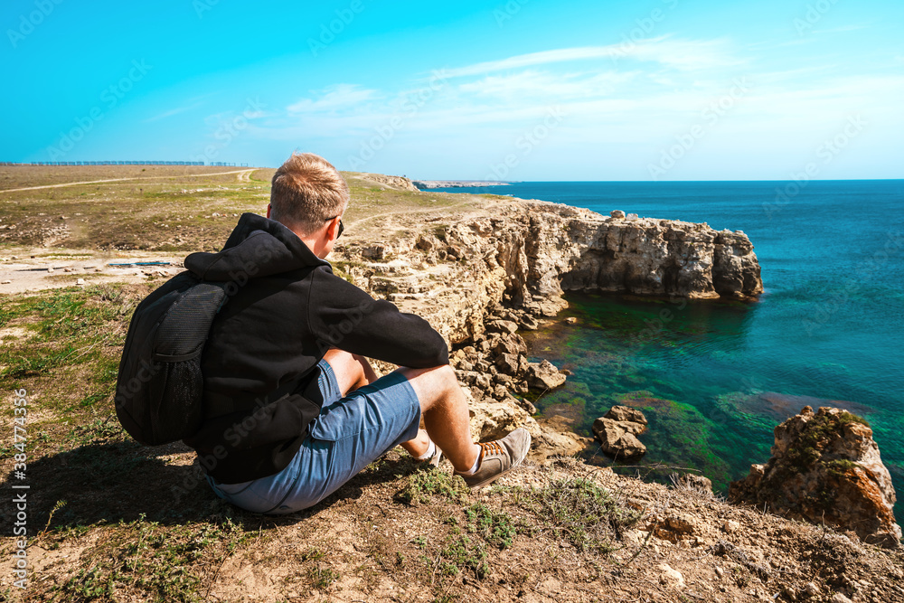 Rear view a young man sits on a high cliff overlooking the rocky cliffs arches on the beach and turquoise sea water on the coast