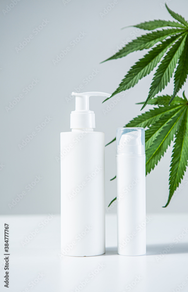 Pair of white mock up blank bottles with CBD natural products and hemp leaves on gray studio background. Pair of white mock up blank bottles with CBD natural products and hemp leaves on gray studio background.
