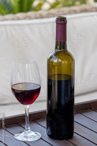 One glass of red wine and an open dark green glass bottle on a wooden serving table near a beige sofa