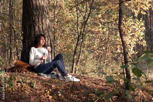 Side view of a traveler young women enjoying a hot drink during hiking sitting on the ground near the tree in park. Girl drinking tea during hike in forest in autumn